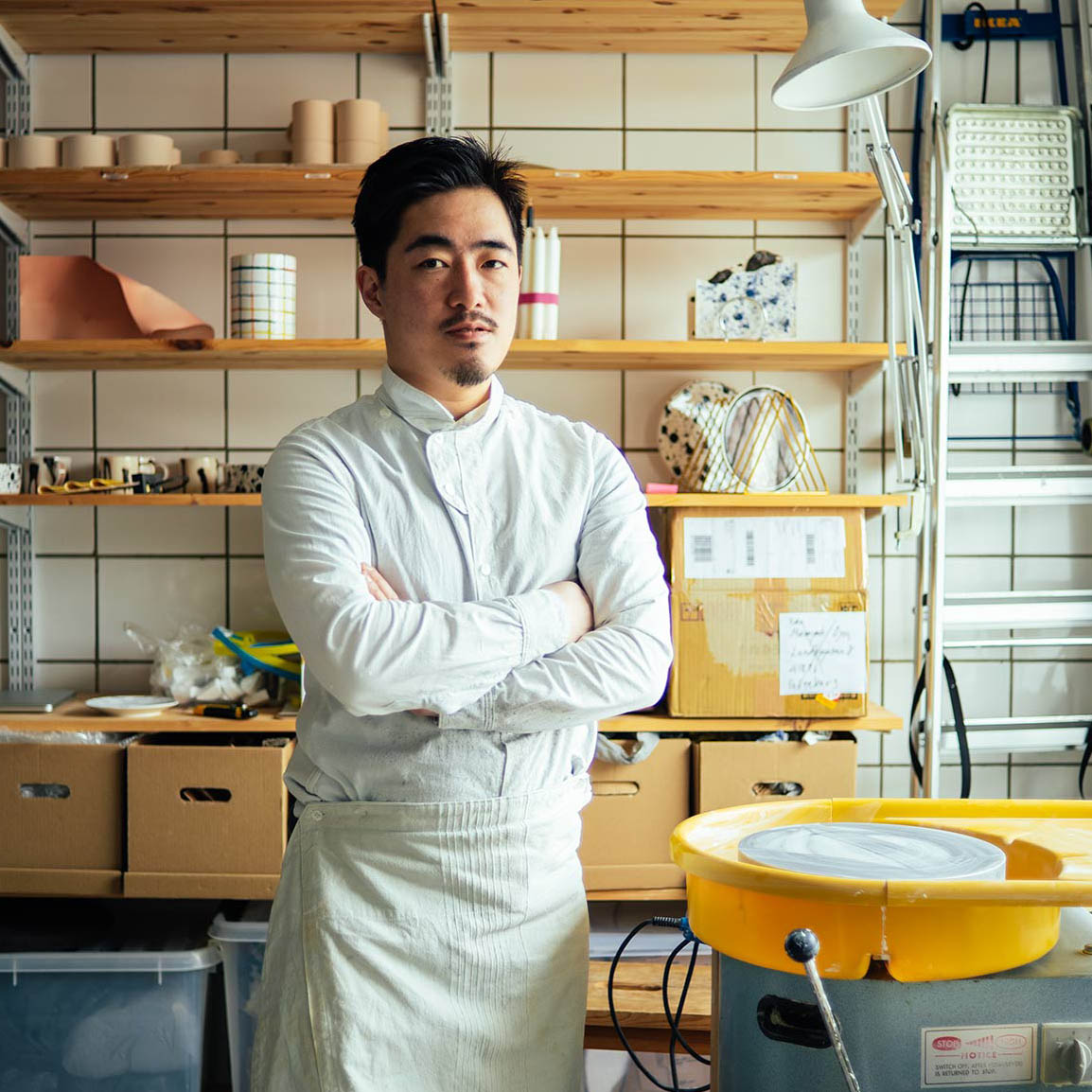 Masayoshi Oya, a ceramic artist from Studio Oyama, standing confidently in his workshop surrounded by shelves with ceramics, tools, and pottery equipment